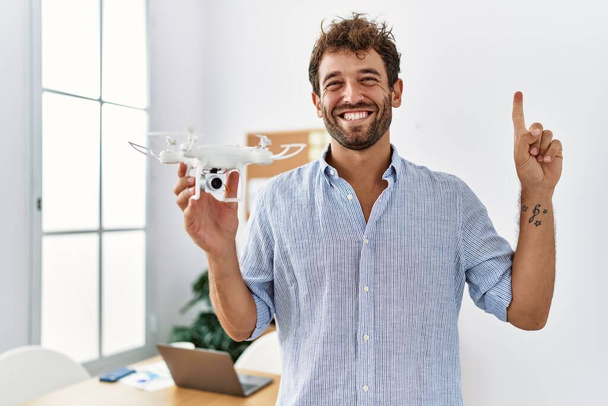 Νεαρός όμορφος άντρας που χρησιμοποιεί τηλεκατευθυνόμενο στο γραφείο του αρχιτέκτονα χαμογελώντας με μια ιδέα ή ερώτηση δείχνοντας το δάχτυλο με χαρούμενο πρόσωπο, νούμερο ένα  - Φωτογραφία, εικόνα