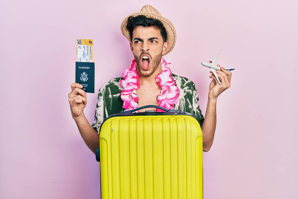 Νεαρός Ισπανός που φοράει καλοκαιρινό στυλ και χαβανέζικο λέι που κρατάει διαβατήριο και αεροπλάνο παιχνίδι θυμωμένος και τρελός που ουρλιάζει απογοητευμένος και έξαλλος, φωνάζοντας από θυμό. οργή και επιθετική αντίληψη.  - Φωτογραφία, εικόνα