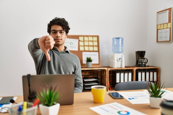 Young hispanic man wearing business style sitting on desk at office looking unhappy and angry showing rejection and negative with thumbs down gesture. bad expression.  - Photo, Image