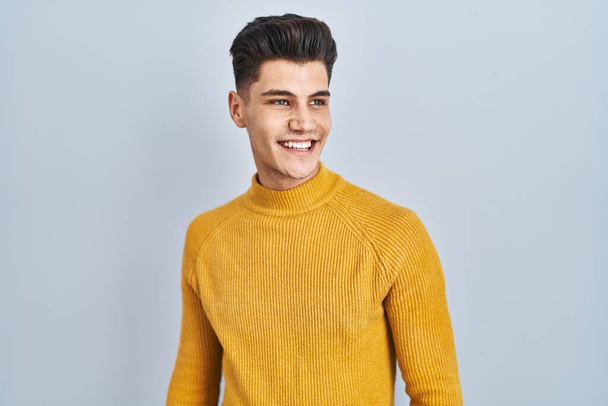 Young hispanic man standing over blue background looking away to side with smile on face, natural expression. laughing confident.  - Foto, imagen