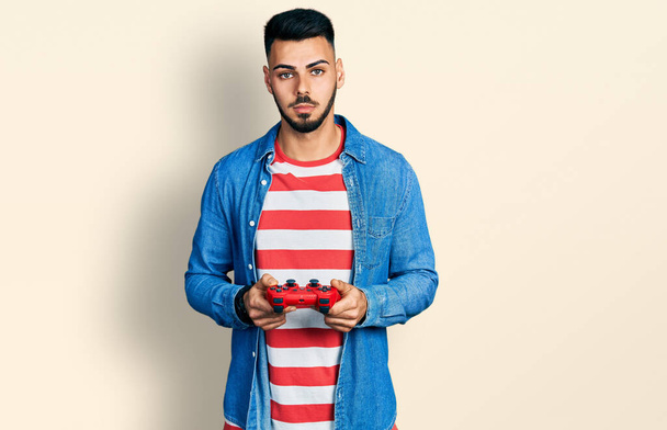Fiatal, szakállas spanyol férfi videojáték-vezérlőt játszik lazán, komoly arckifejezéssel. egyszerű és természetes nézi a kamerát.  - Fotó, kép
