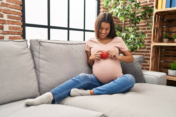Νεαρή Λατίνα γυναίκα έγκυος κρατώντας την καρδιά κάθεται στον καναπέ στο σπίτι - Φωτογραφία, εικόνα