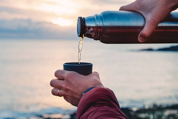 A close-up shot of man pouring hot tea from thermos on the background of the ocean on the sunset - Foto, Imagem