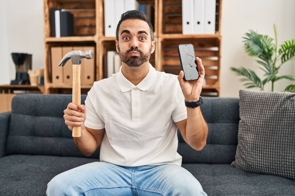 Junger hispanischer Mann mit Bart, Hammer und kaputtem Smartphone, der aufgerissene Bildschirmbacken mit lustigem Gesicht zeigt. Mund mit Luft aufgeblasen, Luft einfangen.  - Foto, Bild
