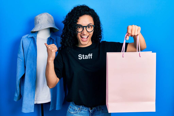 Jonge Spaanse vrouw met krullend haar dragen personeel t shirt houden boodschappentas duim wijzen naar de zijkant glimlachen blij met open mond  - Foto, afbeelding