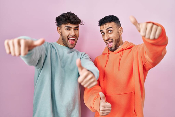 Joven pareja gay hispana de pie sobre fondo rosa aprobando hacer gesto positivo con la mano, pulgares arriba sonriendo y feliz por el éxito. gesto ganador.  - Foto, imagen