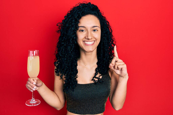 Fiatal, göndör hajú spanyol nő iszik egy pohár pezsgőt, mosolyog egy ötlettel vagy kérdéssel ujjal mutogatva boldog arccal.  - Fotó, kép