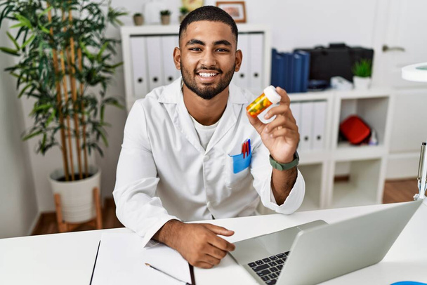 Young indian doctor holding prescription pills looking positive and happy standing and smiling with a confident smile showing teeth  - Valokuva, kuva