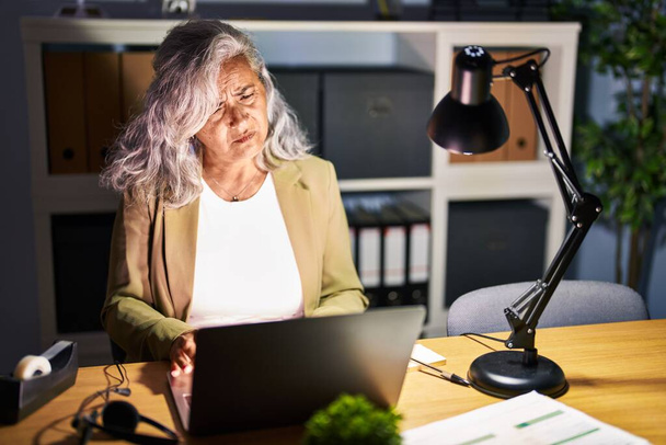 Жінка середнього віку з сірим волоссям, що працює з використанням комп'ютерного ноутбука пізно вночі в депресії і турбується про біду, плаче сердито і боїться. сумний вираз
.  - Фото, зображення