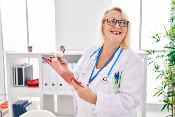 Mujer rubia de mediana edad vistiendo uniforme médico escrito en el documento en la clínica - Foto, Imagen