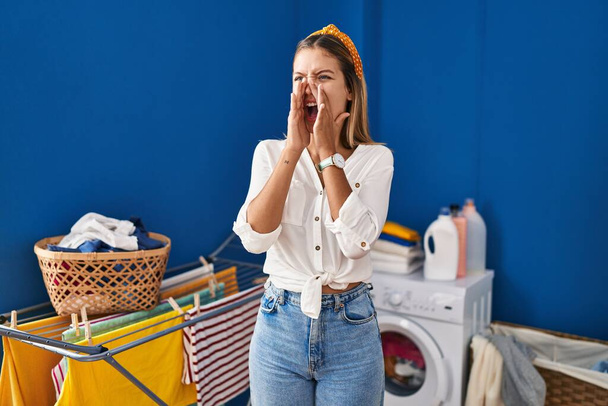 Young blonde woman at laundry room shouting angry out loud with hands over mouth  - 写真・画像