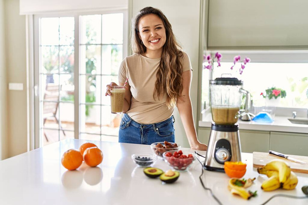 Krásná mladá brunetka žena pití sklenice smoothie v kuchyni vypadá pozitivně a šťastně stojící a usmívá se s sebevědomým úsměvem ukazující zuby  - Fotografie, Obrázek