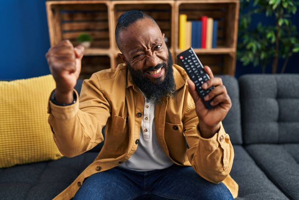 Afrikai amerikai férfi kezében televízió távirányító bosszús és frusztrált kiabál a harag, kiabál őrült a harag és a kéz emelt  - Fotó, kép