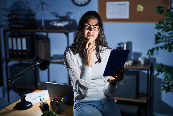 Young brazilian woman using touchpad at night working at the office thinking worried about a question, concerned and nervous with hand on chin  - Foto, Imagem