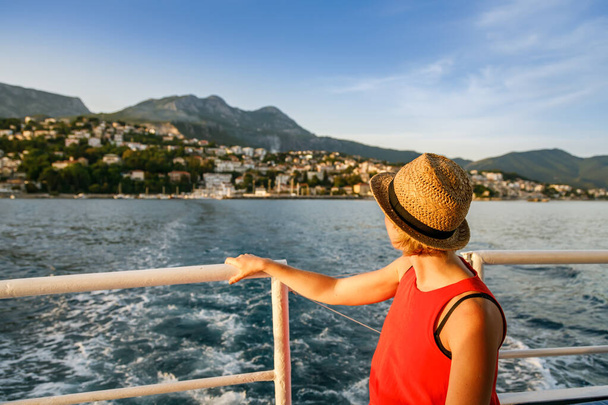Mujer vestida de rojo y sombrero de paja por detrás, mirando a la ciudad de Herceg Novi desde un barco - Kotor Bay, Montenegro - Foto, imagen