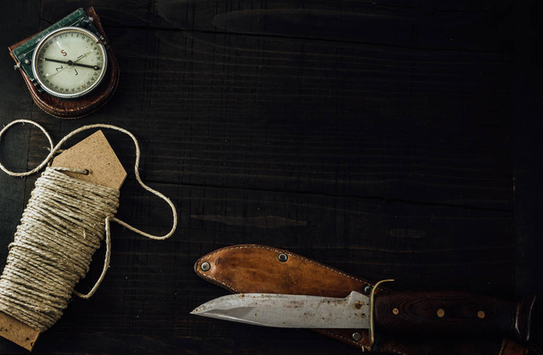 Old rusty hunting bushcraft knife, military compass and a linen rope on the wooden table. Leather cases, copy space, top view, survival concept. - Valokuva, kuva