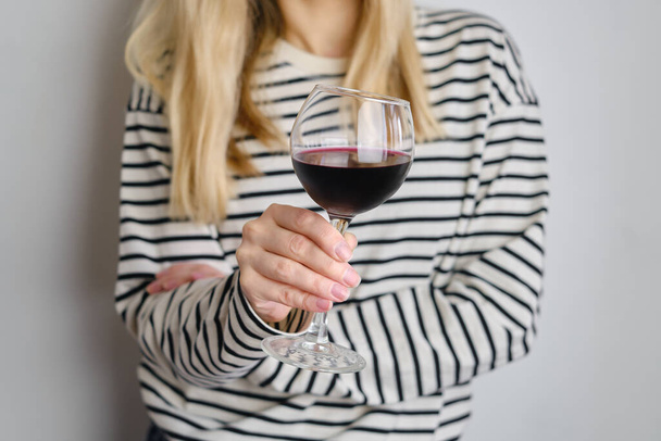 A glass of red wine in the hands of a girl in a striped jacket against the background of a white wall. Wine tasting at the bar. Party, drinks, holidays and celebration concept. Glass close-up - Photo, Image