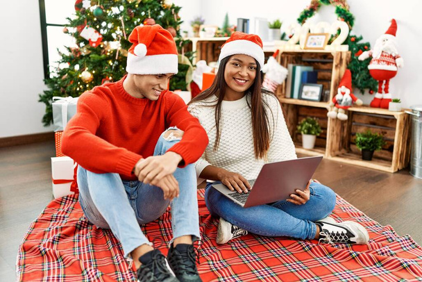Young latin couple smiling happy and using laptop sitting by christmas tree at home. - Fotoğraf, Görsel