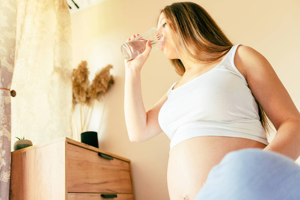 Ciężarna woda pitna. Młoda ciężarna matka pije wodę. Kobieta w ciąży czeka na dziecko. Szklankę wody. Pojęcie macierzyństwa, porodu - Zdjęcie, obraz