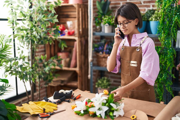 Очаровательная латиноамериканка-флористка, разговаривающая по смартфону, делает букет цветов в цветочном магазине - Фото, изображение