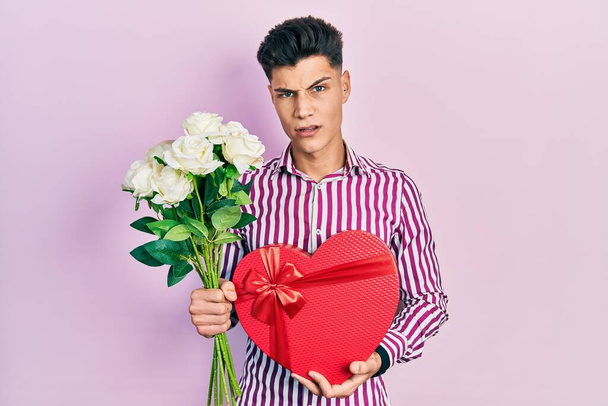 Young hispanic man holding anniversary present and bouquet of flowers in shock face, looking skeptical and sarcastic, surprised with open mouth  - Foto, imagen