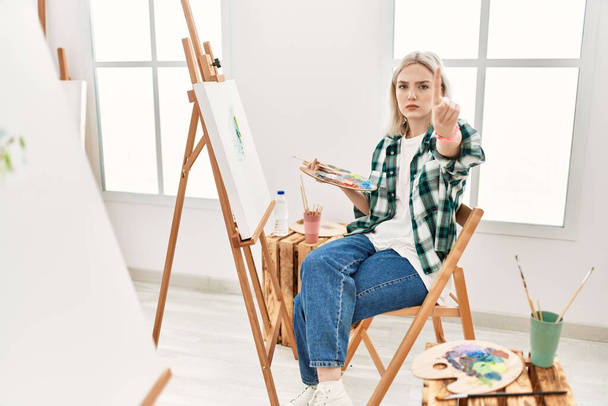 Молодая художница рисует на холсте в художественной студии, показывая пальцем вверх и сердитым выражением лица, не показывая жеста  - Фото, изображение