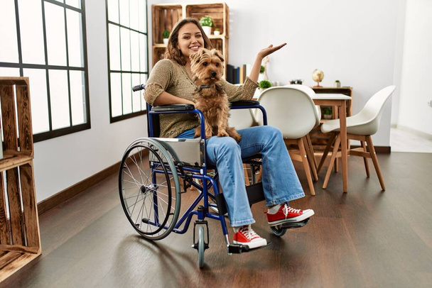 Young hispanic girl sitting on wheelchair at home amazed and smiling to the camera while presenting with hand and pointing with finger.  - Photo, Image