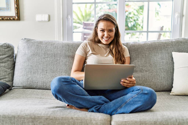Young hispanic girl using laptop sitting on the sofa at home. - Foto, imagen