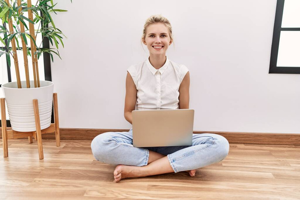 Young blonde woman using computer laptop sitting on the floor at the living room with a happy and cool smile on face. lucky person.  - Foto, Imagem