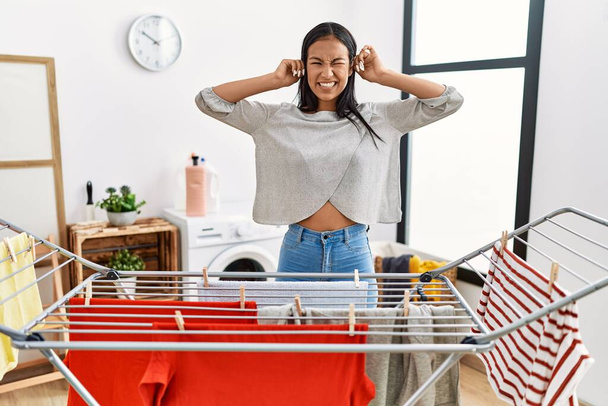Young hispanic woman putting fresh laundry on clothesline covering ears with fingers with annoyed expression for the noise of loud music. deaf concept.  - Photo, Image