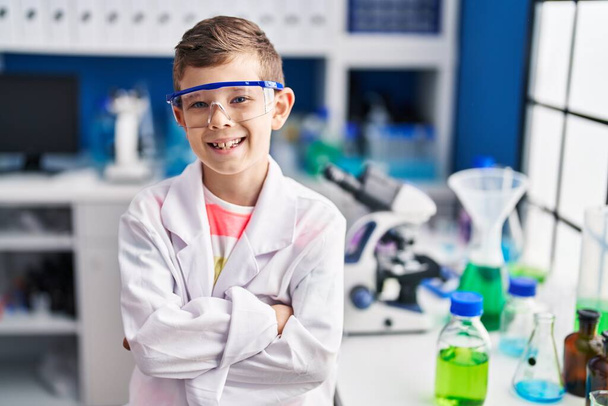 Niño rubio vestido con uniforme científico sentado con los brazos cruzados gesto en el laboratorio - Foto, Imagen