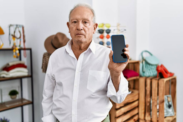 Senior man holding smartphone at retail shop thinking attitude and sober expression looking self confident  - Fotoğraf, Görsel