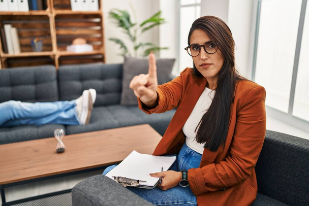 Young hispanic woman working as psychology counselor pointing with finger up and angry expression, showing no gesture  - Фото, изображение