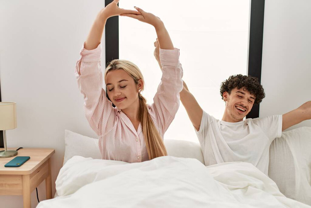 Young beautiful couple yawning and stretching arms sitting on the bed. - Φωτογραφία, εικόνα