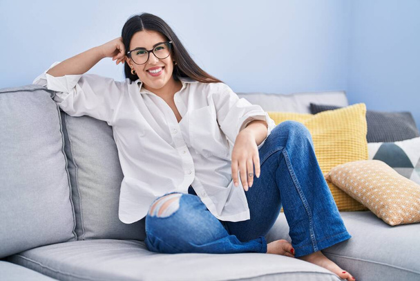 Jovem hispânica sorrindo confiante sentado no sofá em casa - Foto, Imagem