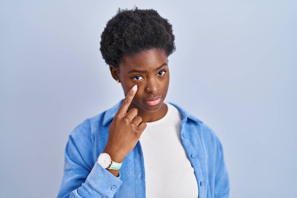 Afrikai amerikai nő áll a kék háttér felé mutató szem figyeli gesztus, gyanús kifejezés  - Fotó, kép