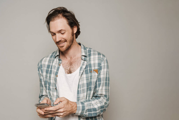 Bristle man dressed in shirt smiling while using cellphone isolated over grey background - Valokuva, kuva