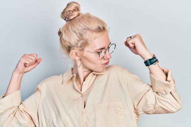 Beautiful caucasian woman with blond hair wearing casual look and glasses showing arms muscles smiling proud. fitness concept.  - 写真・画像