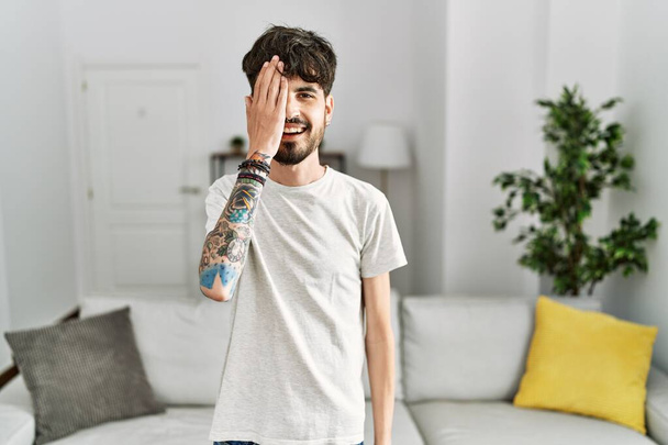 Hispanic man with beard at the living room at home covering one eye with hand, confident smile on face and surprise emotion.  - Foto, afbeelding
