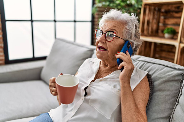 Femme âgée aux cheveux gris buvant du café et parlant sur le smartphone assis sur le canapé à la maison - Photo, image