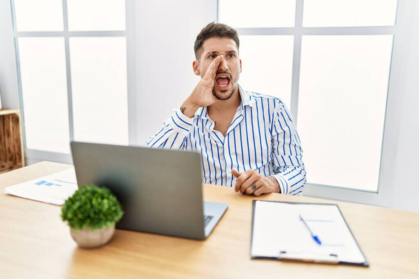 Young handsome man with beard working at the office using computer laptop shouting and screaming loud to side with hand on mouth. communication concept.  - Φωτογραφία, εικόνα