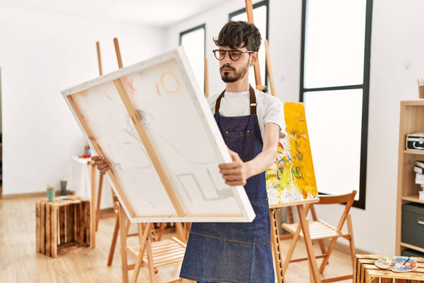 Young hispanic artist man concentrate holding canvas at art studio. - Foto, afbeelding