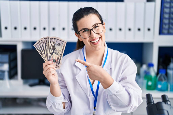 Young brunette woman working at scientist laboratory holding money smiling happy pointing with hand and finger  - 写真・画像