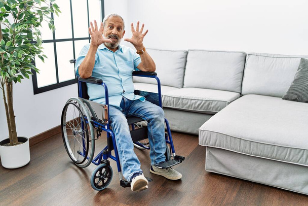 Handsome senior man sitting on wheelchair at the living room showing and pointing up with fingers number ten while smiling confident and happy.  - Fotografie, Obrázek