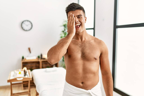 Young hispanic man standing shirtless at spa center covering one eye with hand, confident smile on face and surprise emotion.  - Photo, Image