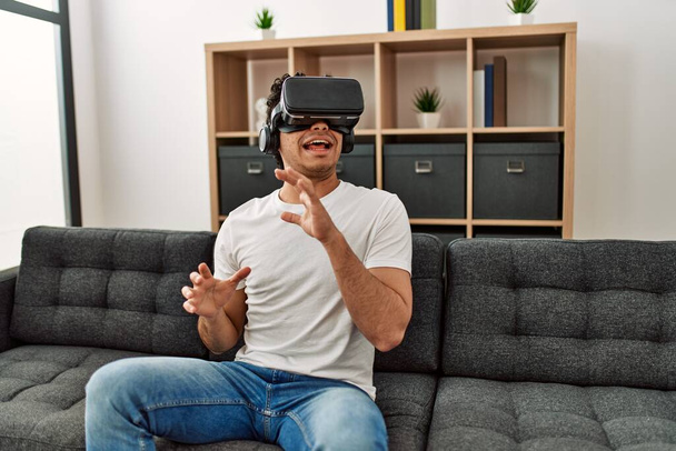 Молодой латиноамериканец боится играть в очки виртуальной реальности, сидя дома на диване. - Фото, изображение