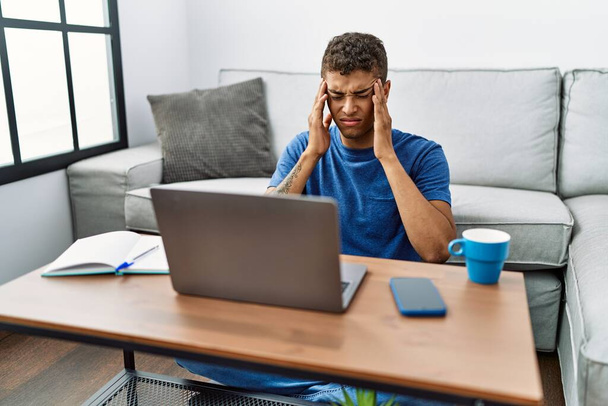 Jeune homme hispanique beau utilisant ordinateur portable assis sur le sol avec la main sur la tête pour la douleur dans la tête parce que le stress. migraine souffrant.  - Photo, image