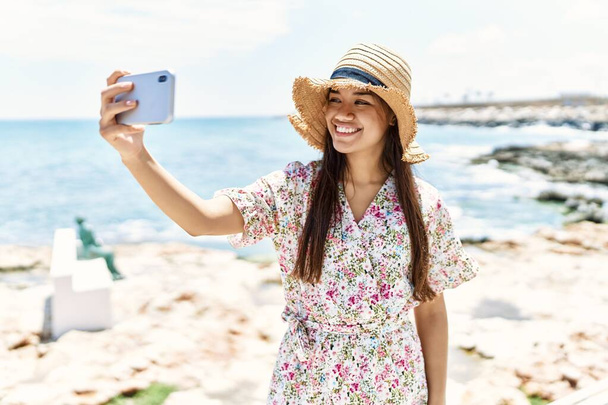 Young latin girl wearing summer hat making selfie by the smartphone at the beach. - Foto, imagen