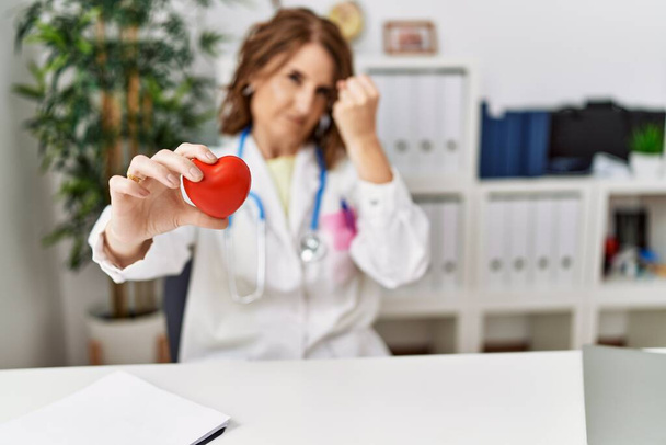 Жінка-лікар середнього віку тримає серце в клініці усміхнений веселий вказує рукою і пальцем вгору на бік
  - Фото, зображення