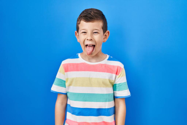 青い背景に立っている若い白人の子供は、面白い表情で幸せに舌を突き出しています。感情の概念.  - 写真・画像